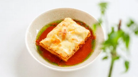 A square of baked lasagna in a bowl with tomato sauce